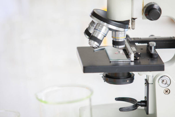 Nahaufnahme von Mikroskoplinse und Probe auf Glasplatte im Laborraum mit buntem Reagenz im Röhrchen und Becher im unscharfen Vordergrund und Hintergrund. - Foto, Bild