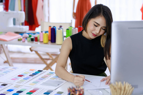 Mujer asiática sonriente modista es vestido de dibujo sentado que tiene que cortar para el cliente en la tienda de sastres y máquinas de coser. Concepto creando nueva colección de ropa - Foto, Imagen