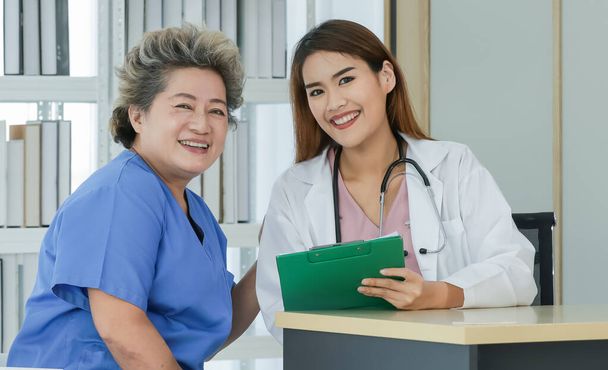 Portrait femme asiatique médecin et patiente âgée portant la robe du patient assis et souriant se sentent à l'aise et heureux en regardant la caméra dans la salle d'examen à l'hôpital le matin. - Photo, image