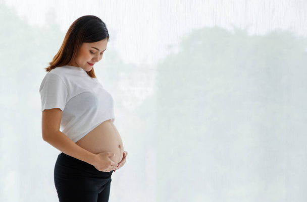 Smiley jong gelukkig zwanger aziatische vrouw dragen terloops staan en aanraken van haar buik met liefde en zorg op zoek naar camera voor studio geschoten tegen witte achtergrond. - Foto, afbeelding