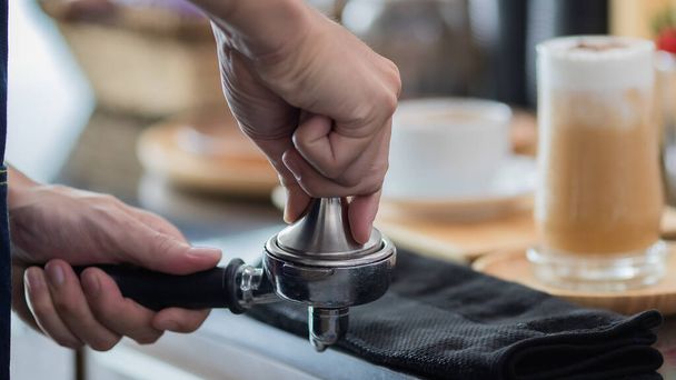 zblízka fotografie mužských rukou držících kovovou manipulaci a portafiltr s kávou v kavárně. Muž barista připravuje pro lisování mleté kávy pro vaření espresso nebo americano v kavárně - Fotografie, Obrázek