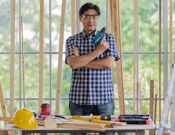 Asijský muž s krátkými černými vlasy inženýr nosit jasné brýle a kostkované košile stojí s úsměvem za pracovním stolem plným domácích zařízení stavební nástroje - Fotografie, Obrázek