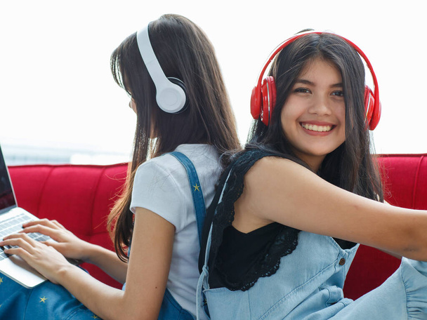 Nahaufnahme eines niedlichen lächelnden jungen thailändisch-türkischen Teenagers, der Rücken an Rücken auf einer roten Couch sitzt und Musik hört. Schwestern mit gemischter Rasse leben zusammen und spielen mit einem Laptop - Foto, Bild