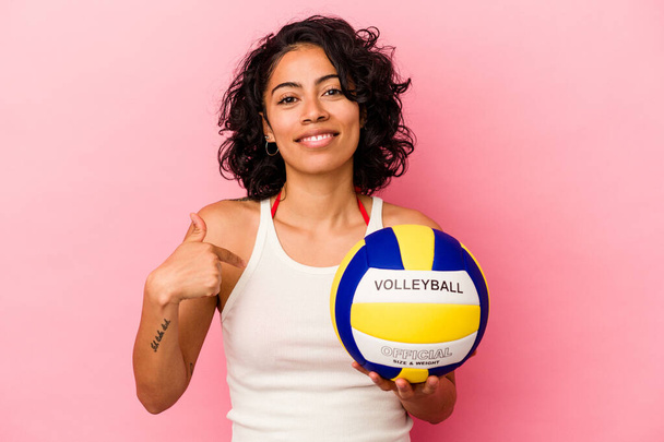 Mujer latina joven sosteniendo una pelota de voleibol aislada sobre fondo rosa persona señalando a mano a un espacio de copia de la camisa, orgulloso y confiado - Foto, imagen