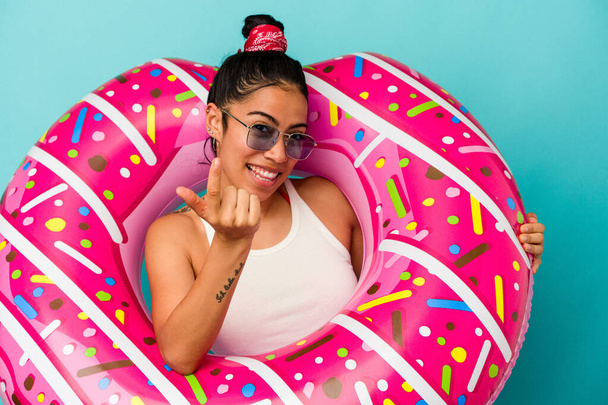 Mulher latina jovem segurando um donut inflável isolado no fundo azul apontando com o dedo para você como se convidando chegar mais perto. - Foto, Imagem