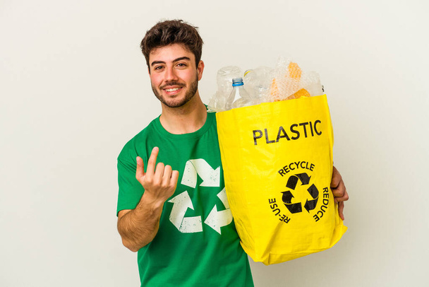 Young caucasian man recycling plastic isolated on white background pointing with finger at you as if inviting come closer. - Photo, Image
