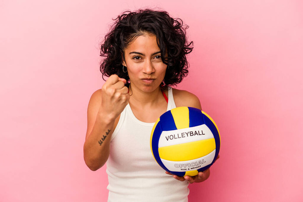 Mujer latina joven sosteniendo una bola de volea aislada sobre fondo rosa mostrando puño a cámara, expresión facial agresiva. - Foto, Imagen