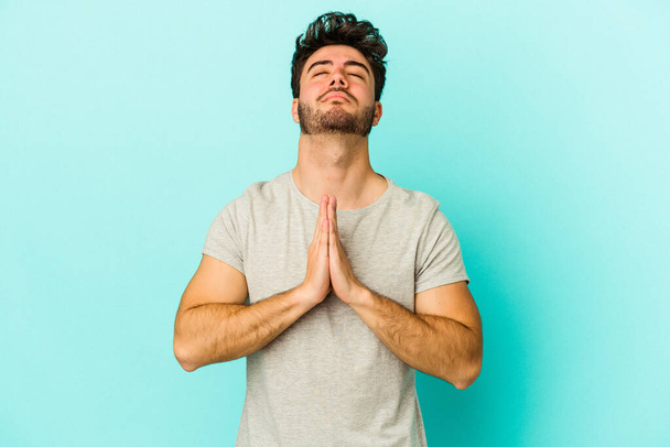 Young caucasian man isolated on blue background holding hands in pray near mouth, feels confident. - Zdjęcie, obraz