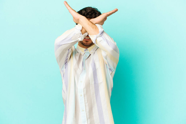 Молодой кавказский человек, изолированный на синем фоне, держащий две руки скрещенными, концепция отрицания. - Фото, изображение