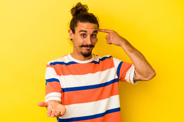 Young caucasian man with long hair isolated on yellow background holding and showing a product on hand. - Foto, afbeelding