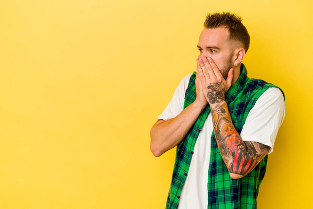 Νεαρός καυκάσιος με τατουάζ απομονωμένος σε κίτρινο φόντο στοχαστικός κοιτάζοντας σε ένα χώρο αντιγραφής που καλύπτει το στόμα με το χέρι. - Φωτογραφία, εικόνα