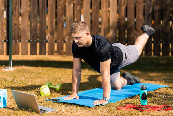 The young man goes in for sports outside. Cheerful sportsman with blond hair makes the lunges on lawn in garden, next to there is a laptop with online training - Photo, Image