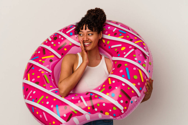 Junge gemischte Rasse Frau hält einen aufblasbaren Air Donut isoliert auf weißem Hintergrund sagt eine geheime heiße Bremsnachrichten und schaut zur Seite - Foto, Bild