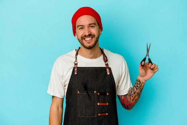 Young tattooed barber man isolated on blue background happy, smiling and cheerful. - Photo, Image