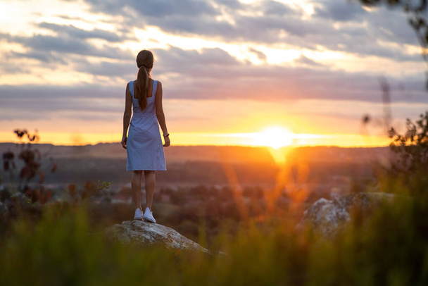 Ciemna sylwetka młodej kobiety w letniej sukience stojącej na świeżym powietrzu z widokiem na naturę o zachodzie słońca. - Zdjęcie, obraz
