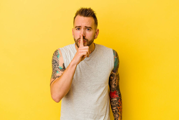 Joven hombre caucásico tatuado aislado sobre fondo amarillo manteniendo un secreto o pidiendo silencio. - Foto, imagen