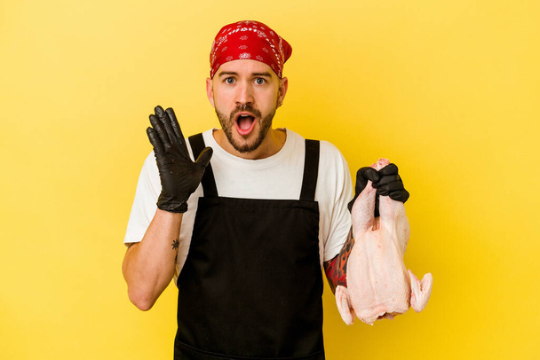 Fiatal tetovált batcher kaukázusi férfi kezében egy csirke elszigetelt sárga háttér meglepett és sokkolt. - Fotó, kép