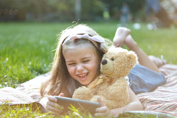 Klein kind meisje op zoek in haar mobiele telefoon samen met haar favoriete teddybeer speelgoed buiten in de zomer park. - Foto, afbeelding
