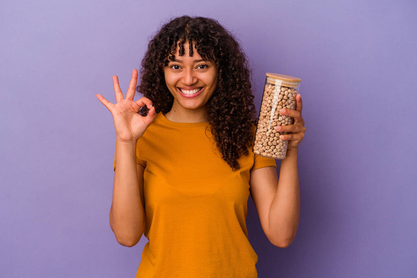 Giovane donna brasiliana in possesso di una bottiglia di ceci isolato su sfondo viola allegro e fiducioso mostrando gesto ok. - Foto, immagini
