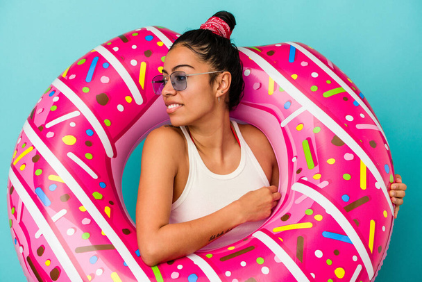 Mulher latina jovem segurando um donut inflável isolado no fundo azul olha para o lado sorridente, alegre e agradável. - Foto, Imagem