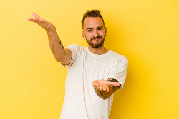 Young tattooed caucasian man isolated on yellow background makes scale with arms, feels happy and confident. - Photo, Image