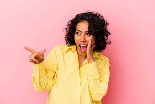 Young curly latin woman isolated on pink background saying a gossip, pointing to side reporting something. - Foto, Imagen