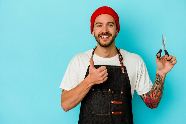 Young tattooed barber man isolated on blue background smiling and raising thumb up - Photo, Image