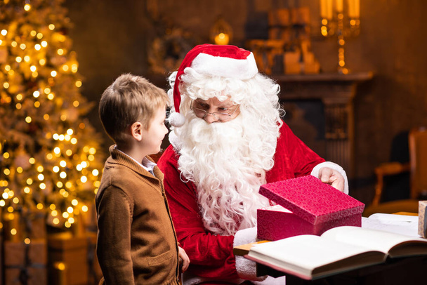Santa Claus and little boy. Cheerful Santa is working while sitting at the table. Fireplace and Christmas Tree in the background. Christmas concept. - Фото, зображення