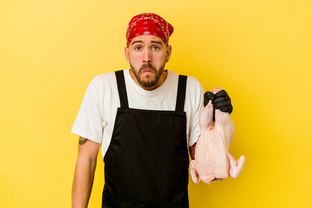 Nuori tatuoitu paimen valkoihoinen mies pitämässä kanaa eristettynä keltaisella taustalla kohauttaa olkapäitä ja avoimet silmät sekaisin. - Valokuva, kuva