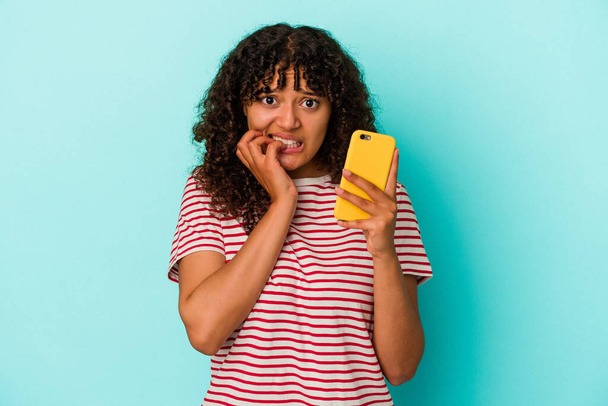 Молодая женщина смешанной расы держит мобильный телефон изолирован на синем фоне грызет ногти, нервный и очень тревожный. - Фото, изображение