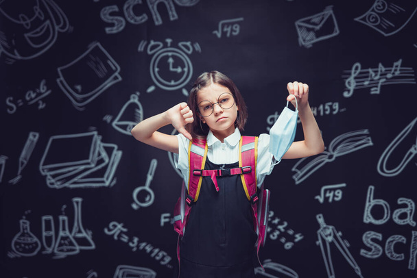 Caucasian sad schoolgirl showing thumb down holding medical mask in hand. Back to school concept  - Photo, Image