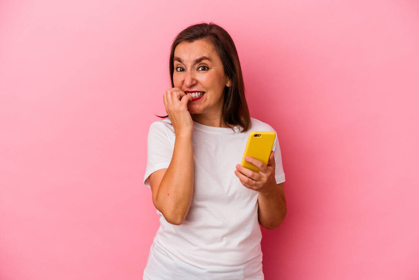 Середньовічна біла жінка тримає мобільний телефон ізольовано на рожевому фоні кусаючи нігті, нервує і дуже тривожна
. - Фото, зображення