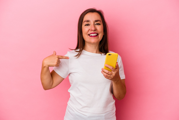 Middle age caucasian woman holding mobile phone isolated on pink background person pointing by hand to a shirt copy space, proud and confident - Photo, Image