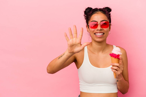 Junge lateinische Frau hält ein Eis auf rosa Hintergrund isoliert lächelnd fröhlich zeigt Nummer fünf mit den Fingern. - Foto, Bild