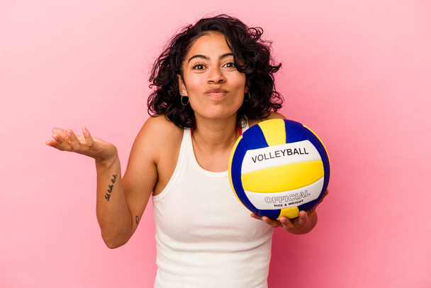 Mujer latina joven sosteniendo una bola de volea aislada sobre fondo rosa encoge hombros y los ojos abiertos confundidos. - Foto, imagen