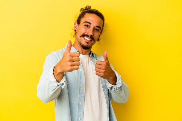 Young caucasian man with long hair isolated on yellow background raising both thumbs up, smiling and confident. - Photo, image