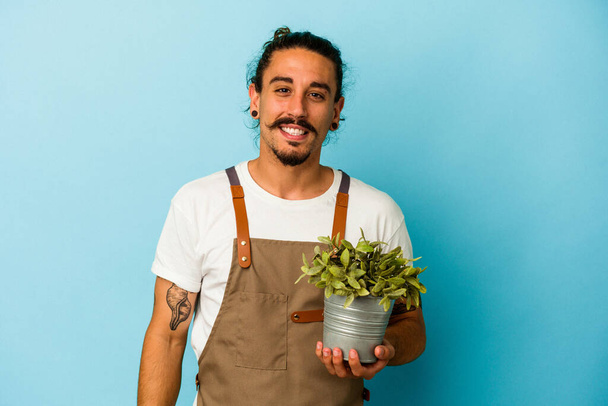 Молодий садівник білий чоловік тримає рослину ізольовано на синьому фоні щасливий, усміхнений і веселий
. - Фото, зображення