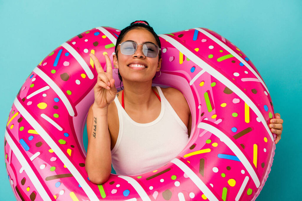 Mulher latina jovem segurando um donut inflável isolado no fundo azul mostrando o número dois com os dedos. - Foto, Imagem