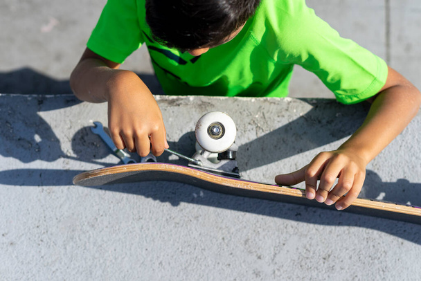 Bovenaanzicht van een jongen die het wiel van zijn skateboard repareert nadat hij het op een zonnige dag in het skateboard park heeft gebruikt. - Foto, afbeelding