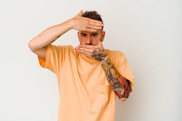 Young tattooed caucasian man isolated on white background blink at the camera through fingers, embarrassed covering face. - Photo, Image
