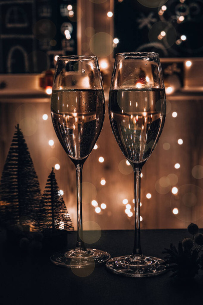 Два бокала шампанского с игристыми бокалами. Темный фон с желтым светобоком. Рождественские игрушки на столе. Зимние каникулы - Фото, изображение