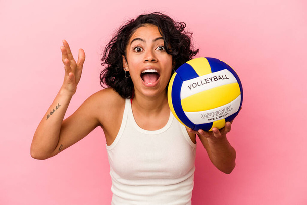 Mujer latina joven sosteniendo una pelota de voleibol aislada sobre fondo rosa recibiendo una agradable sorpresa, excitada y levantando las manos. - Foto, imagen