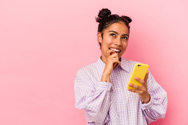 Junge lateinamerikanische Frau hält ein Handy isoliert auf rosa Hintergrund und denkt entspannt über etwas nach, das einen Kopierraum betrachtet. - Foto, Bild