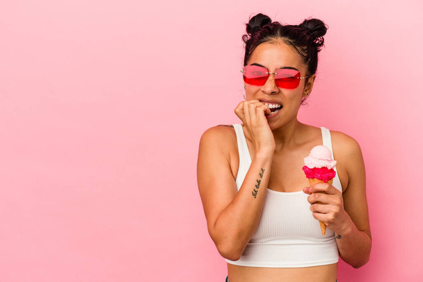 Νεαρή Λατίνα γυναίκα που κρατάει ένα παγωτό απομονωμένο σε ροζ φόντο δαγκώνοντας νύχια, νευρική και πολύ αγχωμένη. - Φωτογραφία, εικόνα