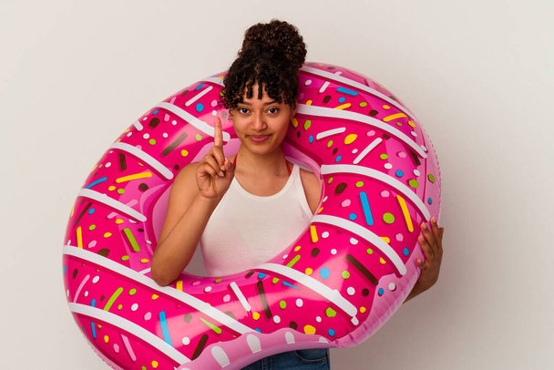 Молодая женщина смешанной расы держит надувной воздушный пончик изолирован на белом фоне показывая номер один с пальцем. - Фото, изображение