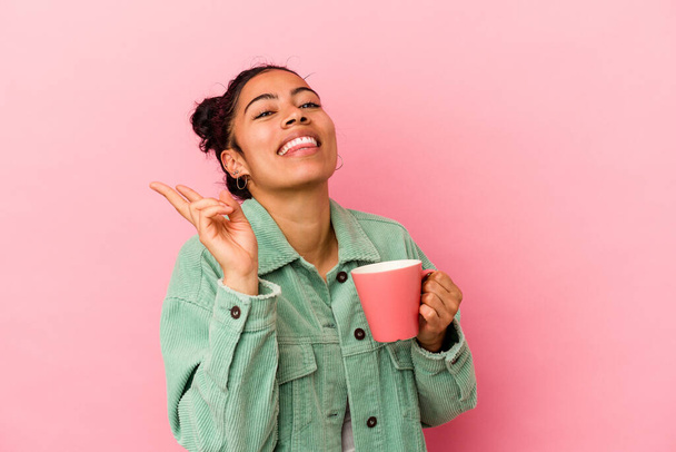Giovane donna latina che tiene una tazza isolata su sfondo rosa gioiosa e spensierata mostrando un simbolo di pace con le dita. - Foto, immagini