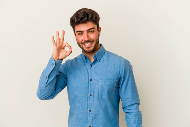Junger kaukasischer Mann, isoliert auf weißem Hintergrund, zwinkert und hält eine Okay-Geste mit der Hand. - Foto, Bild