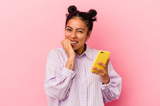 Молода латинка тримає мобільний телефон ізольовано на рожевому фоні кусає нігті, нервує і дуже тривожно
. - Фото, зображення