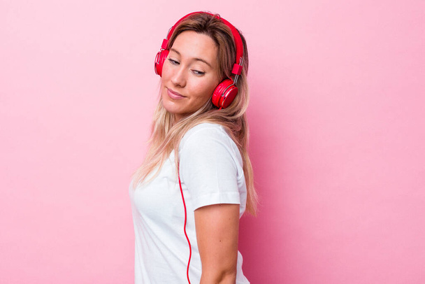 Young australian woman listening music isolated on pink background looks aside smiling, cheerful and pleasant. - Photo, Image