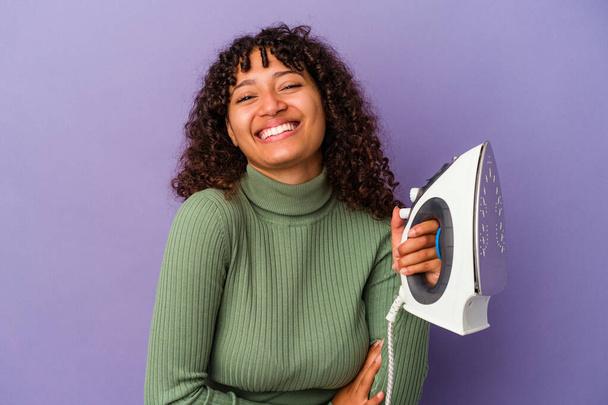Young mixed race woman holding an iron isolated on purple background laughing and having fun. - Photo, Image